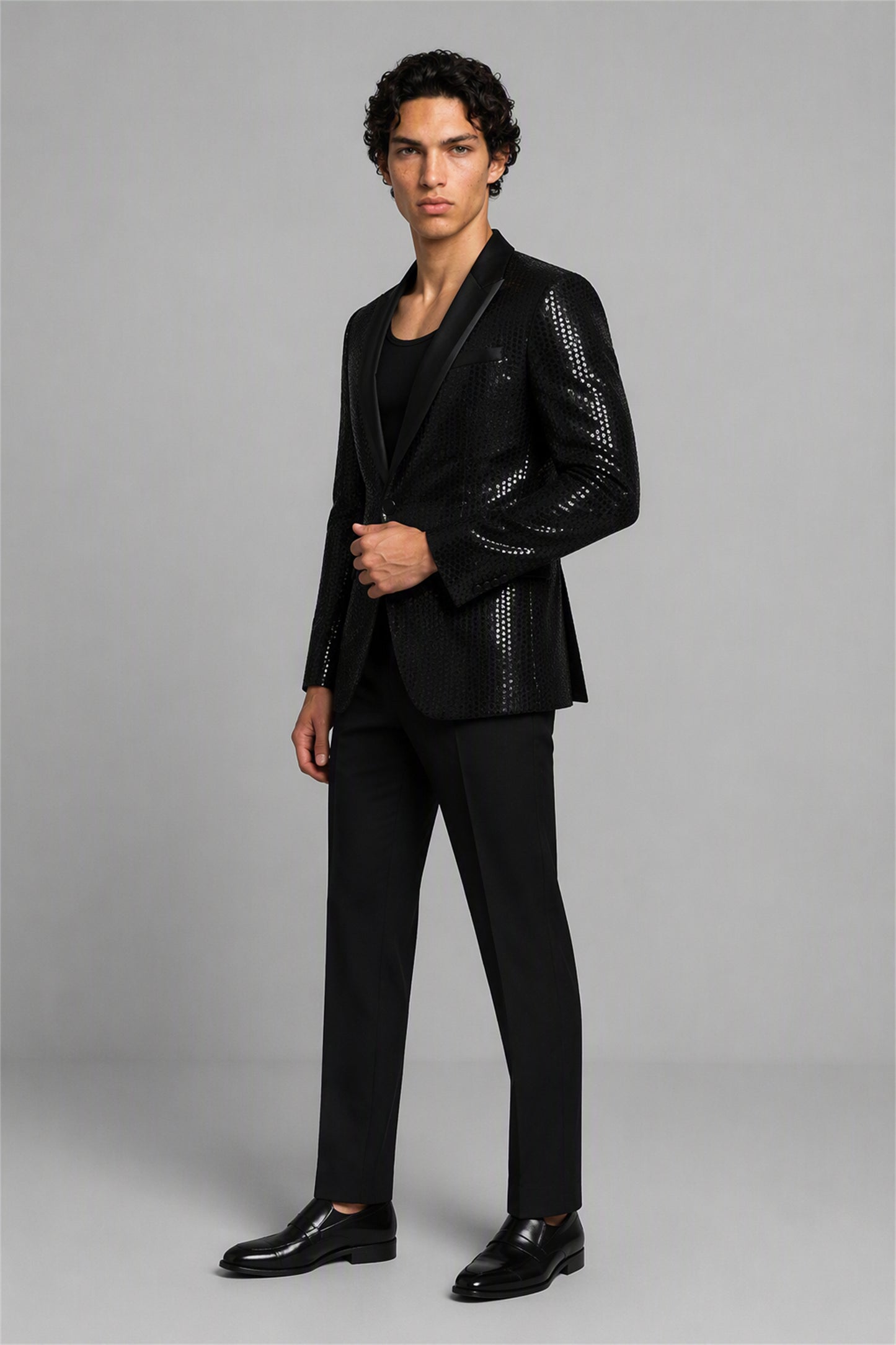 Savile Black Sequin Tuxedo Jacket – Three-quarter front standing view, hand on button showing sequin embellishment and peak lapel contrast